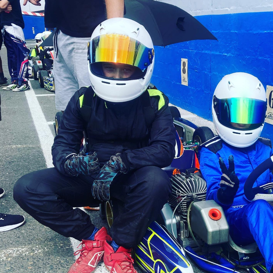 foto di due piloti di kart dopo la corsa parcheggiati in pista. A destra, il pilota ,ancora seduto sul suo kart, ha una tuta blu e casco bianco con visiera blu e riflessi verdi. Accanto a lui, a sinistra, l'altro pilota appoggiato al kart del compagno. Indossa una tuta nera e ha un casco bianco con visiera blu e riflessi arancioni. Quello a sinistra fa il gesto del pollice in su mentre l'altro fa il segno "vittoria" con l'indice e il medio. In sfondo, si vedono altri piloti in riposo come loro.